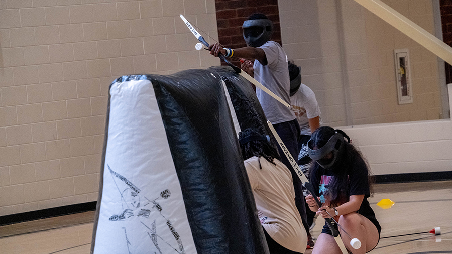 Group of students in a gym wearing safety goggles, using long sticks to hit a small white ball toward a padded obstacle.
