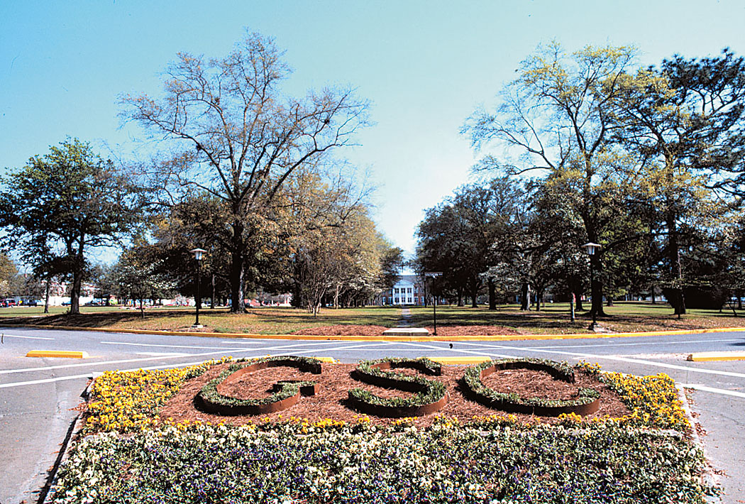 Roundabout with a flower-bed sculpture spelling 'GSO' in green hedges, surrounded by yellow flowers and trees, with a building in the background.”] }? Wait need correct JSON array without extra. Let's produce proper.