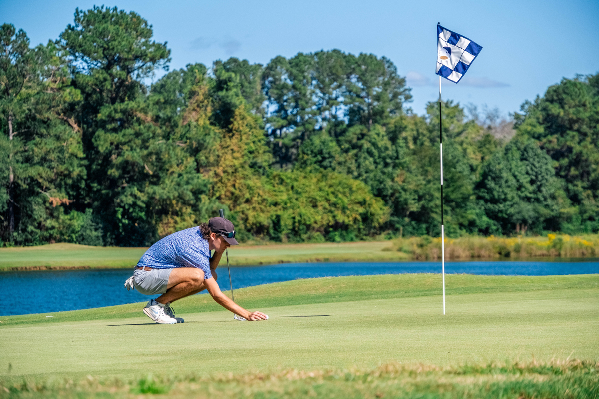 Golfer kneeling on the green, lining up a putt near the hole with a flag in the background by a water hazard