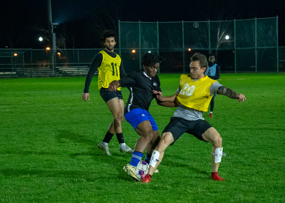 Two players in yellow bibs and one in black contest the ball during a night soccer match.