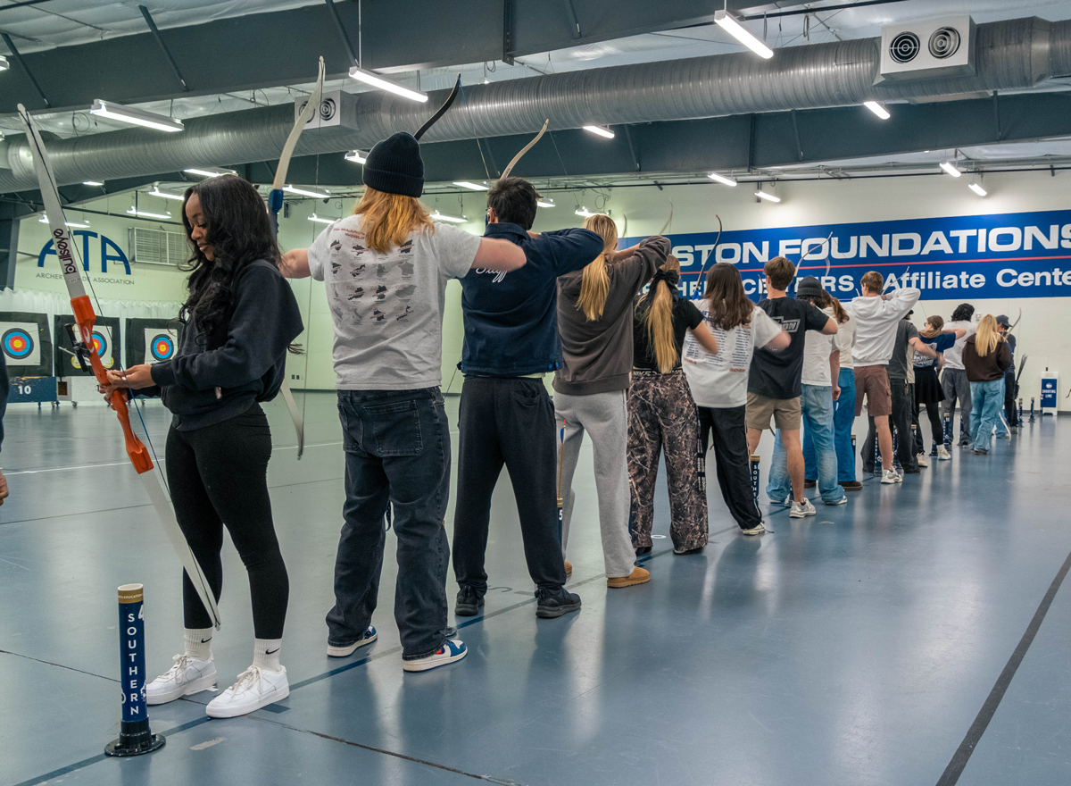 Line of people in an indoor archery range, each archer drawing a bow, targets visible at the back of the room.