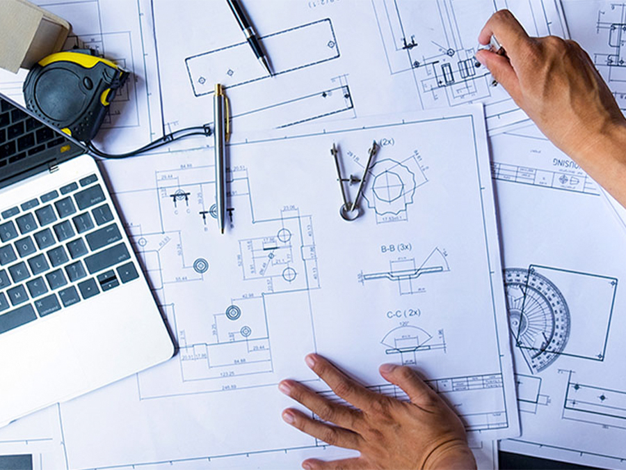 Hands working on technical blueprints with engineering tools and a laptop.