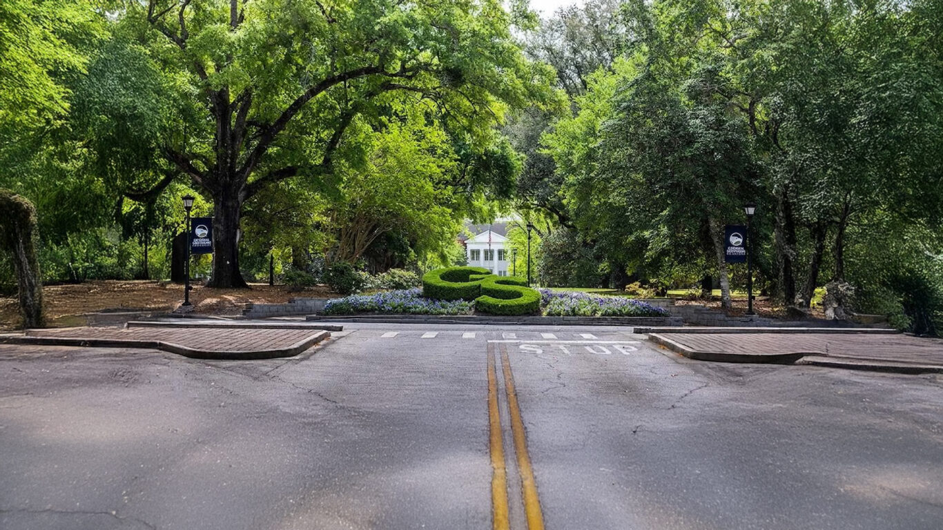 Tree-lined street leading to a manicured circular hedge and a white gazebo beyond, framed by lush green trees and lampposts with banners on either side.